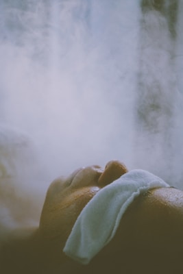 A person is lying down with a white cloth or towel placed over their head, surrounded by mist or steam. The scene suggests a relaxing or therapeutic environment.