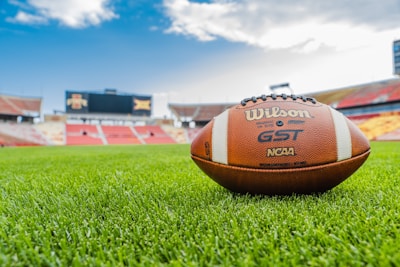 A football lies on a lush, well-manicured field in a stadium setting. The background features empty stands and a large scoreboard under a blue sky with clouds.