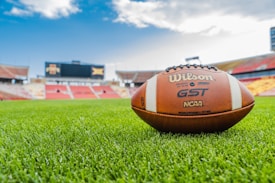 A football lies on a lush, well-manicured field in a stadium setting. The background features empty stands and a large scoreboard under a blue sky with clouds.