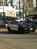 A police vehicle is driving along a city street. The background features a building with various signs, including a large advertisement for a donair shop. The street is busy with other vehicles and pedestrians. The scene is urban with modern buildings and trees lining the street.
