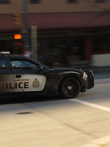 A police car from Vancouver is in motion on a city street. The car is a black and white vehicle with the word 'POLICE' and an emblem visible on the side. The background is blurred, indicating speed, and includes urban elements like an intersection and traffic lights.