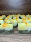 Multiple handmade soap bars are arranged in rows on a wooden surface. Each soap bar has a gradient appearance, transitioning from dark green at the bottom to yellow and white at the top. Small orange bits are sprinkled on the top of each soap bar. The brand name 'Himeriez' is embossed on each of the bars.
