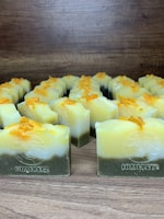 Multiple handmade soap bars are arranged in rows on a wooden surface. Each soap bar has a gradient appearance, transitioning from dark green at the bottom to yellow and white at the top. Small orange bits are sprinkled on the top of each soap bar. The brand name 'Himeriez' is embossed on each of the bars.