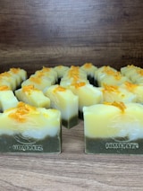 Multiple handmade soap bars are arranged in rows on a wooden surface. Each soap bar has a gradient appearance, transitioning from dark green at the bottom to yellow and white at the top. Small orange bits are sprinkled on the top of each soap bar. The brand name 'Himeriez' is embossed on each of the bars.