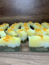 Multiple handmade soap bars are arranged in rows on a wooden surface. Each soap bar has a gradient appearance, transitioning from dark green at the bottom to yellow and white at the top. Small orange bits are sprinkled on the top of each soap bar. The brand name 'Himeriez' is embossed on each of the bars.