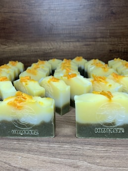 Multiple handmade soap bars are arranged in rows on a wooden surface. Each soap bar has a gradient appearance, transitioning from dark green at the bottom to yellow and white at the top. Small orange bits are sprinkled on the top of each soap bar. The brand name 'Himeriez' is embossed on each of the bars.