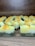 Multiple handmade soap bars are arranged in rows on a wooden surface. Each soap bar has a gradient appearance, transitioning from dark green at the bottom to yellow and white at the top. Small orange bits are sprinkled on the top of each soap bar. The brand name 'Himeriez' is embossed on each of the bars.