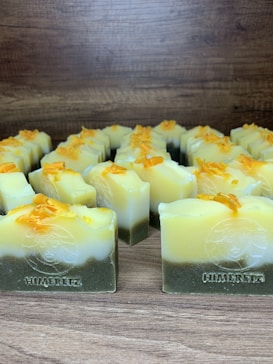 Multiple handmade soap bars are arranged in rows on a wooden surface. Each soap bar has a gradient appearance, transitioning from dark green at the bottom to yellow and white at the top. Small orange bits are sprinkled on the top of each soap bar. The brand name 'Himeriez' is embossed on each of the bars.