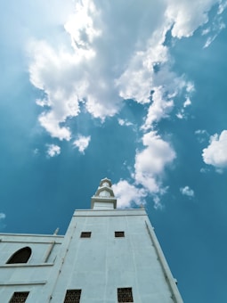 A tall white building with a tower extending towards a bright blue sky filled with scattered white clouds. The architecture has several rectangular and arched windows.