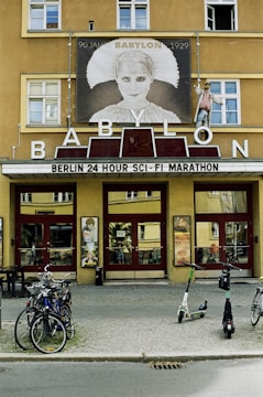 A historic cinema building with a large sign displaying 'BABYLON' in bold letters. Above the entrance is a poster of a character from the classic film 'Metropolis' with the text '90 Jahre BABYLON 1929'. The marquee advertises a 'Berlin 24 Hour Sci-Fi Marathon'. Bicycles and electric scooters are parked in front of the entrance, and a window cleaner is working on the building facade.