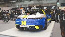A high-performance sports car with police markings is displayed at an indoor automotive event. The car features a blue and yellow color scheme with the word 'Polizei' on the rear, and it sits on a white platform. Surrounding the vehicle, there are people attending the event, some of whom are wearing masks. Various banners and automotive equipment are visible in the background, creating an industrial atmosphere.