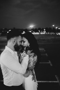 A black and white photograph of a couple embracing closely in a dimly lit area, possibly a parking lot at night. The woman is wearing a sleeveless dress and has visible tattoos on her arm, and the man is dressed in a button-down shirt. Their foreheads are touching, conveying intimacy and tenderness.
