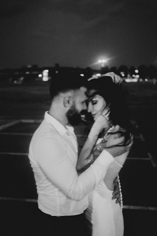 A black and white photograph of a couple embracing closely, conveying a sense of intimacy and affection. The background appears to be a dimly lit outdoor setting at night, with blurred lights and silhouettes in the distance.