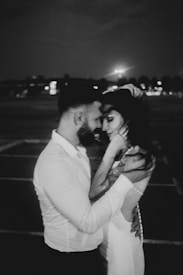 A black and white photograph of a couple embracing closely, conveying a sense of intimacy and affection. The background appears to be a dimly lit outdoor setting at night, with blurred lights and silhouettes in the distance.