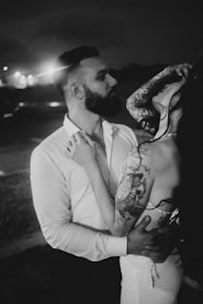A black and white image featuring an intimate moment between two people, a man and a woman. The man has a beard and is wearing a white shirt, while the woman, who has long hair and a visible arm tattoo, is holding onto the man's shirt and tilting her head back. The background is dark and slightly blurred, with some lights visible in the distance.