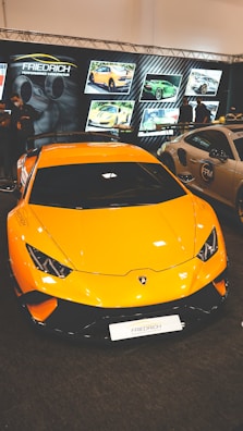 A vibrant yellow sports car with sleek design, prominently displayed in a showroom or exhibition setting. Several screens in the background showcase different images of high-performance cars. Lighting is focused on the cars, highlighting their features.