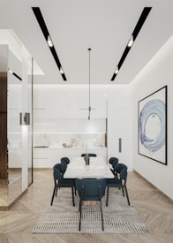 A modern kitchen-dining area featuring a sleek white design. A long dining table with a white tabletop and dark blue chairs occupies the center. Above the table hangs a minimalist black pendant light. The kitchen in the background has white cabinetry and a white marble backsplash. The light wood parquet flooring adds warmth to the space. A large abstract painting with predominantly blue hues adorns the wall to the right. Recessed ceiling lights provide additional illumination.