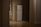 A dimly lit hallway with wooden floors features a white refrigerator situated against a wall. There is a radiator next to a window on the left side, and the space has a minimalistic appearance with neutral-colored walls.