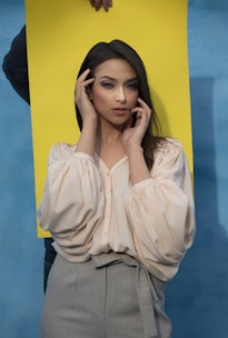 A woman with long dark hair is wearing a light-colored blouse and grey trousers. She is posing with her hands framing her face, standing in front of a blue wall and a bright yellow backdrop held by someone.
