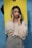 A woman with long dark hair is wearing a light-colored blouse and grey trousers. She is posing with her hands framing her face, standing in front of a blue wall and a bright yellow backdrop held by someone.