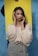 A woman with long dark hair is wearing a light-colored blouse and grey trousers. She is posing with her hands framing her face, standing in front of a blue wall and a bright yellow backdrop held by someone.