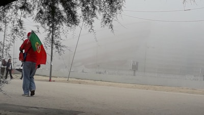 A person wearing a red jacket and jeans is walking on a foggy street, carrying a Portuguese flag. The scene is obscured by dense fog, with faint outlines of a large building and a fence in the background. There are a few other people in the distance.