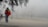 A person wearing a red jacket and jeans is walking on a foggy street, carrying a Portuguese flag. The scene is obscured by dense fog, with faint outlines of a large building and a fence in the background. There are a few other people in the distance.