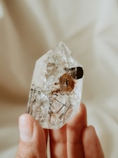 A hand is holding a clear crystal with dark inclusions inside. The background is out of focus, featuring soft, neutral colors that enhance the transparency and texture of the crystal.