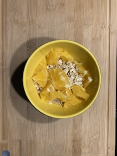 A yellow bowl containing a mixture of oats and orange segments, placed on a wooden surface. The arrangement is simple and the ingredients appear fresh.