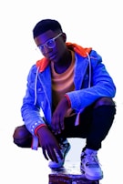 A young person is crouching, wearing a light blue denim jacket with an orange hooded sweatshirt underneath. They have short hair and are wearing glasses. Their outfit includes ripped black jeans and white sneakers with a black and white pattern. The lighting gives a cool, modern feel with shades of blue and purple.