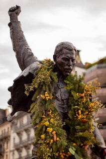 A bronze statue of a man with his fist raised in the air, adorned with green and yellow floral garlands. The background shows buildings with ornate architecture, indicating an urban setting.