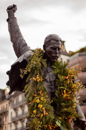 A bronze statue of a man with his fist raised in the air, adorned with green and yellow floral garlands. The background shows buildings with ornate architecture, indicating an urban setting.