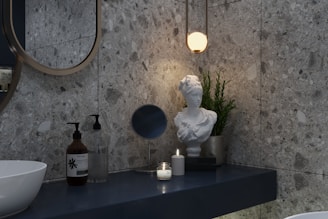 A modern bathroom scene featuring a marble-textured wall and a sleek, minimalist countertop. On the counter are two pump bottles, a round mirror, a lit candle, and a white marble bust placed next to a green potted plant. Above them hangs a warm, glowing pendant light.