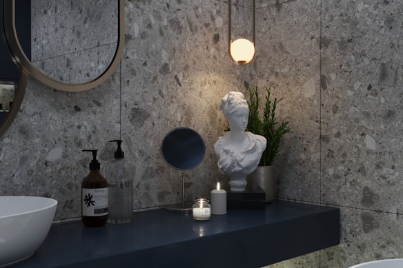 A modern bathroom scene featuring a marble-textured wall and a sleek, minimalist countertop. On the counter are two pump bottles, a round mirror, a lit candle, and a white marble bust placed next to a green potted plant. Above them hangs a warm, glowing pendant light.