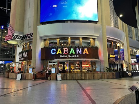 A restaurant with a wooden facade located in a modern urban setting. Above the entrance, a bright and colorful sign spells out the name 'CABANA' in large letters. The exterior is adorned with decorative plants and lights. There are a few informational boards placed outside, and a digital screen with various languages welcomes visitors. The area is well-lit and features modern architectural elements with large columns.
