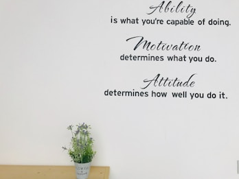 Inspirational text is displayed on the wall, featuring phrases about ability, motivation, and attitude. Below the quotes, a small plant with purple flowers is placed on a wooden surface.