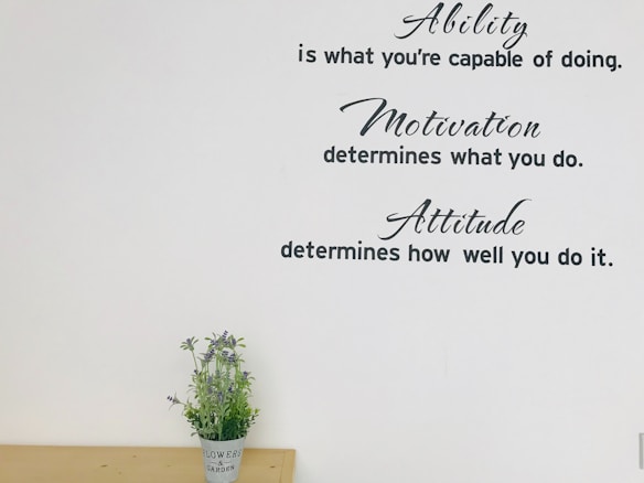 Inspirational text is displayed on the wall, featuring phrases about ability, motivation, and attitude. Below the quotes, a small plant with purple flowers is placed on a wooden surface.