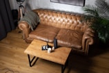 A cozy living room setting features a brown leather tufted sofa with a green throw blanket draped over one arm. In front of the sofa is a wooden coffee table with a candle, a book, and a reed diffuser placed on top. A tall potted plant adds a touch of greenery to the right side of the scene, and a tripod floor lamp stands in the corner, contributing to the room's warm and inviting atmosphere.