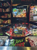 A pinball machine surrounded by books, featuring vibrant and detailed artwork with a horror theme. The machine displays colorful graphics with characters and scenery, including text elements like 'Scared Stiff' and 'Six Tales of Terror,' set in an atmospheric, dimly-lit room with bookshelves in the background.
