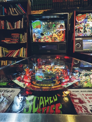 A pinball machine surrounded by books, featuring vibrant and detailed artwork with a horror theme. The machine displays colorful graphics with characters and scenery, including text elements like 'Scared Stiff' and 'Six Tales of Terror,' set in an atmospheric, dimly-lit room with bookshelves in the background.