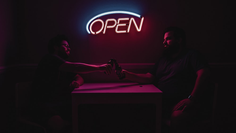 a man and a woman sitting at a table in front of a neon sign