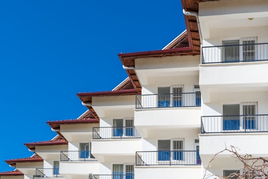 A modern apartment building with multiple floors, white exterior walls, and dark brown roof edges. Each unit has a balcony with black railing, and large windows that reflect the blue sky.