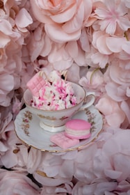 A delicate teacup filled with pink and white whipped topping and garnished with pink candies sits on a matching saucer. Surrounding the teacup are soft pink floral petals creating a romantic and elegant setting.