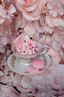 A delicate teacup filled with pink and white whipped topping and garnished with pink candies sits on a matching saucer. Surrounding the teacup are soft pink floral petals creating a romantic and elegant setting.