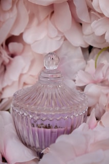 A decorative glass jar with a ribbed design and a round lid is surrounded by delicate light pink flower petals. The jar contains a purple substance, likely a type of wax or gel. The soft petals create an elegant and feminine backdrop.