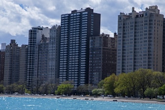A row of tall residential buildings stands along a waterfront, with clear water in the foreground and a partially cloudy sky overhead. The buildings exhibit a mix of architectural styles, featuring both modern and older designs. Trees line the waterfront, hinting at a park or promenade area.