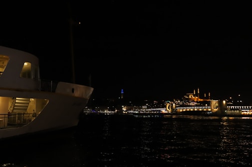 A nighttime scene featuring a large, illuminated ferry on the left side, with visible stairs and passengers. In the background, a well-lit bridge spans across the water, leading to a mosque with glowing minarets and domes, set against a dark sky.