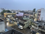 A densely packed urban neighborhood is visible with a mix of old and new buildings. The streets are wet, suggesting recent rain, and a few people are walking along the street while a white van is parked nearby. Overhead, power lines crisscross the scene, and streetlights cast a soft glow. In the background, palm trees and various rooftops are visible.