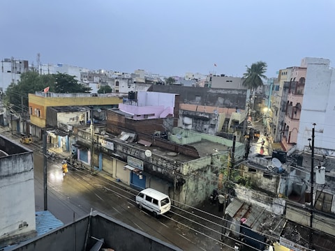 A densely packed urban neighborhood is visible with a mix of old and new buildings. The streets are wet, suggesting recent rain, and a few people are walking along the street while a white van is parked nearby. Overhead, power lines crisscross the scene, and streetlights cast a soft glow. In the background, palm trees and various rooftops are visible.