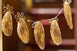 Several banana slices are hanging on a line using unique flower-shaped clips. The bananas appear to be dried, with a golden-brown color and visible dark spots along the length. The background is softly blurred, adding focus to the hanging bananas.